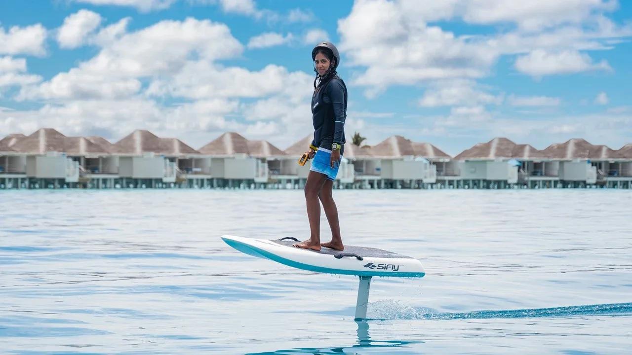 A guest riding a SiFly e-foil board across the lagoon, overwater villas lined up along the horizon behind her
