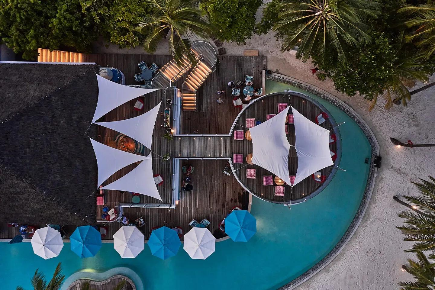 An aerial view of Beach Club at dusk, its sail-shaped canopies, circular day-bed lounge and pool lit for the evening