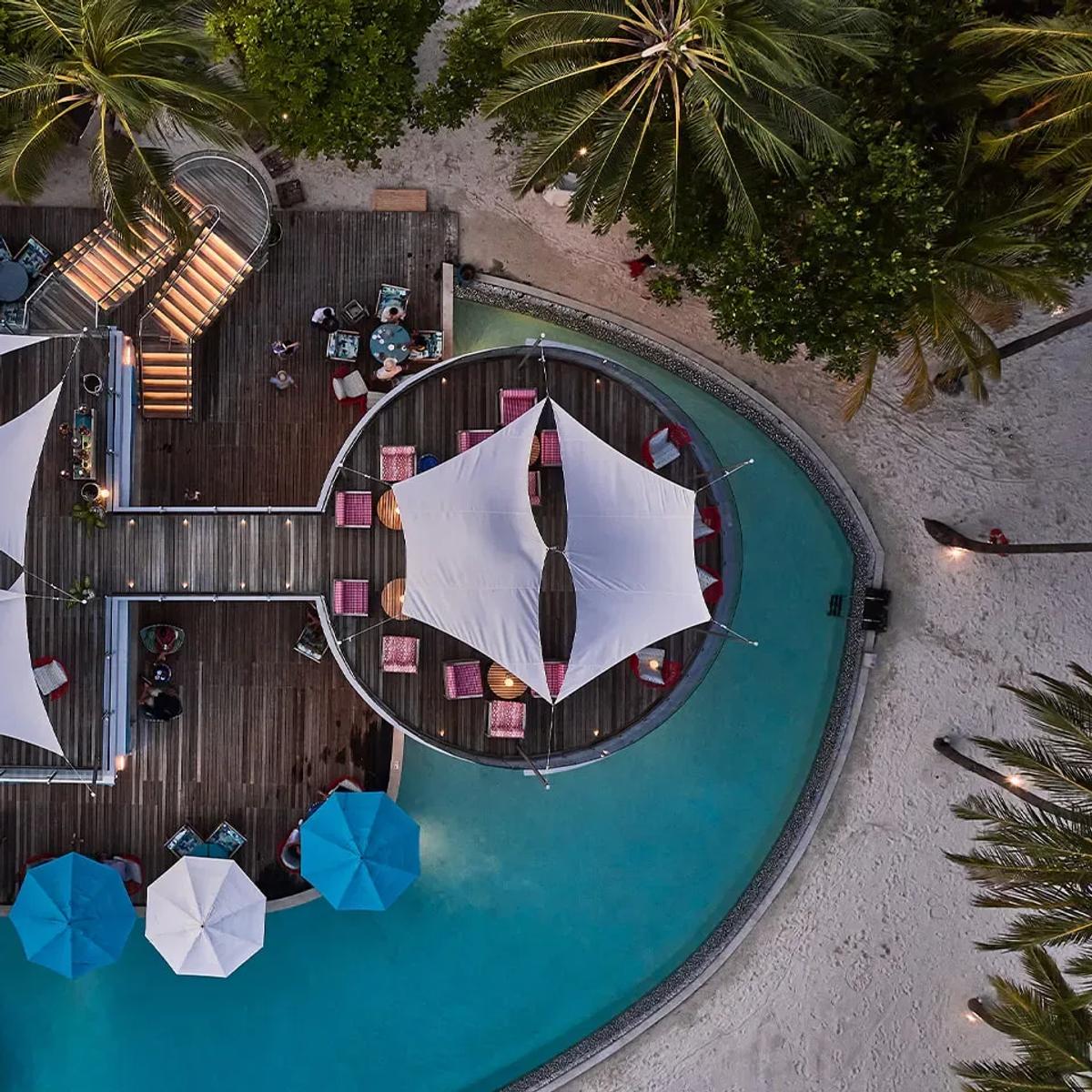 An aerial view of Beach Club at dusk, its sail-shaped canopies, circular day-bed lounge and pool lit for the evening