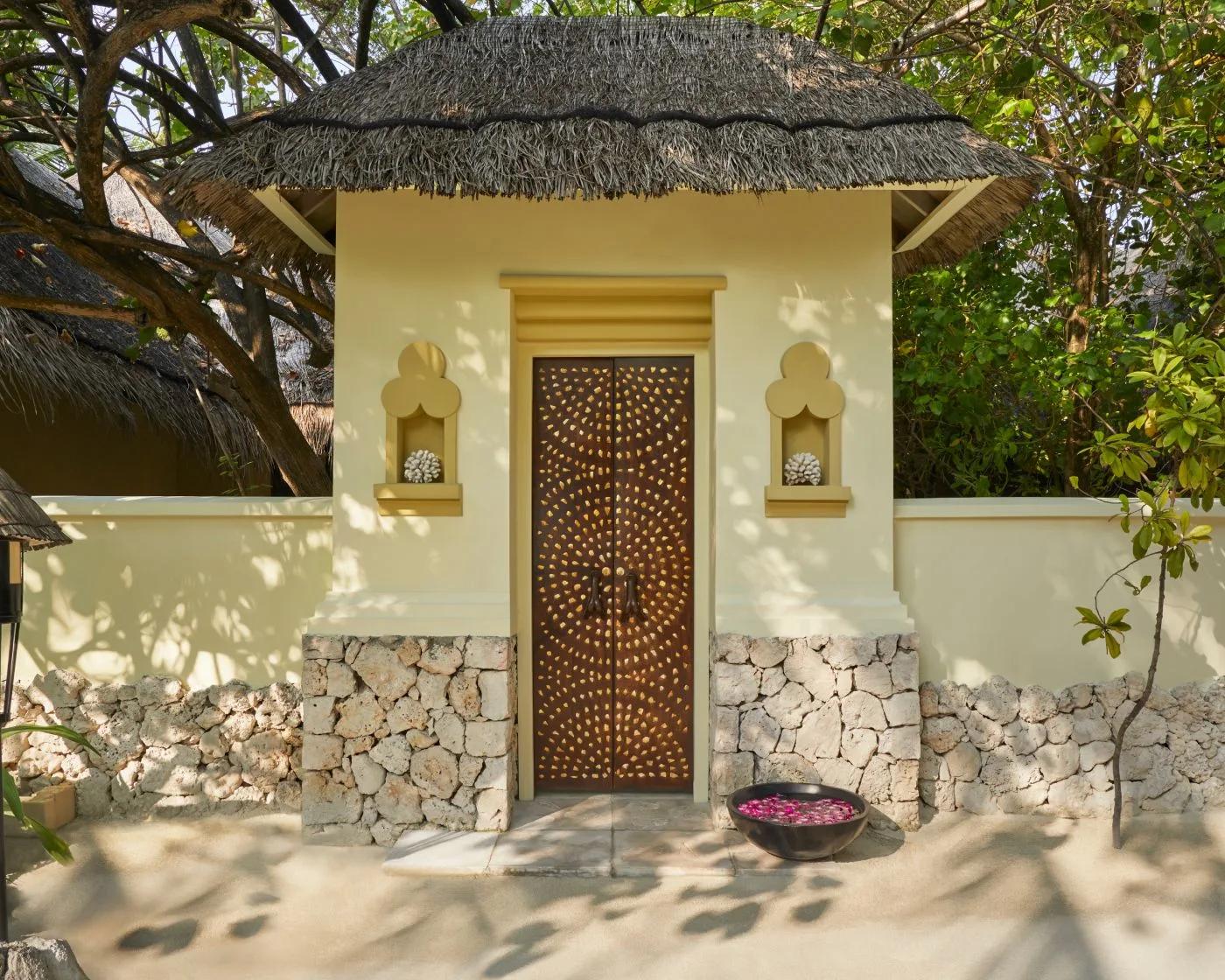 A carved wooden door set into a yellow, thatched-roof pavilion, coral pieces displayed in wall niches either side