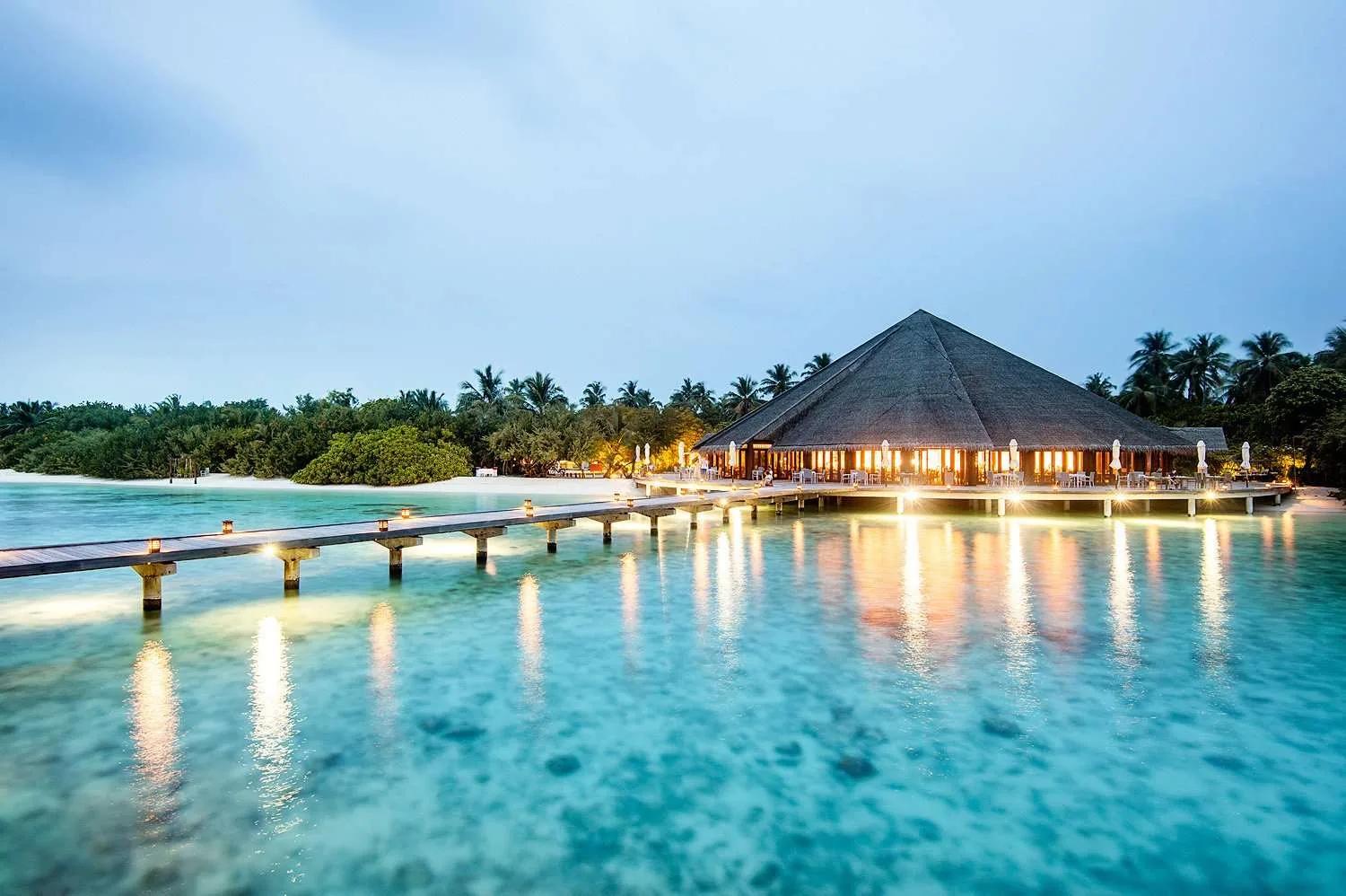 Matheefaru Restaurant lit at dusk, its jetty and thatched roof over the lagoon, Hideaway Beach Resort & Spa