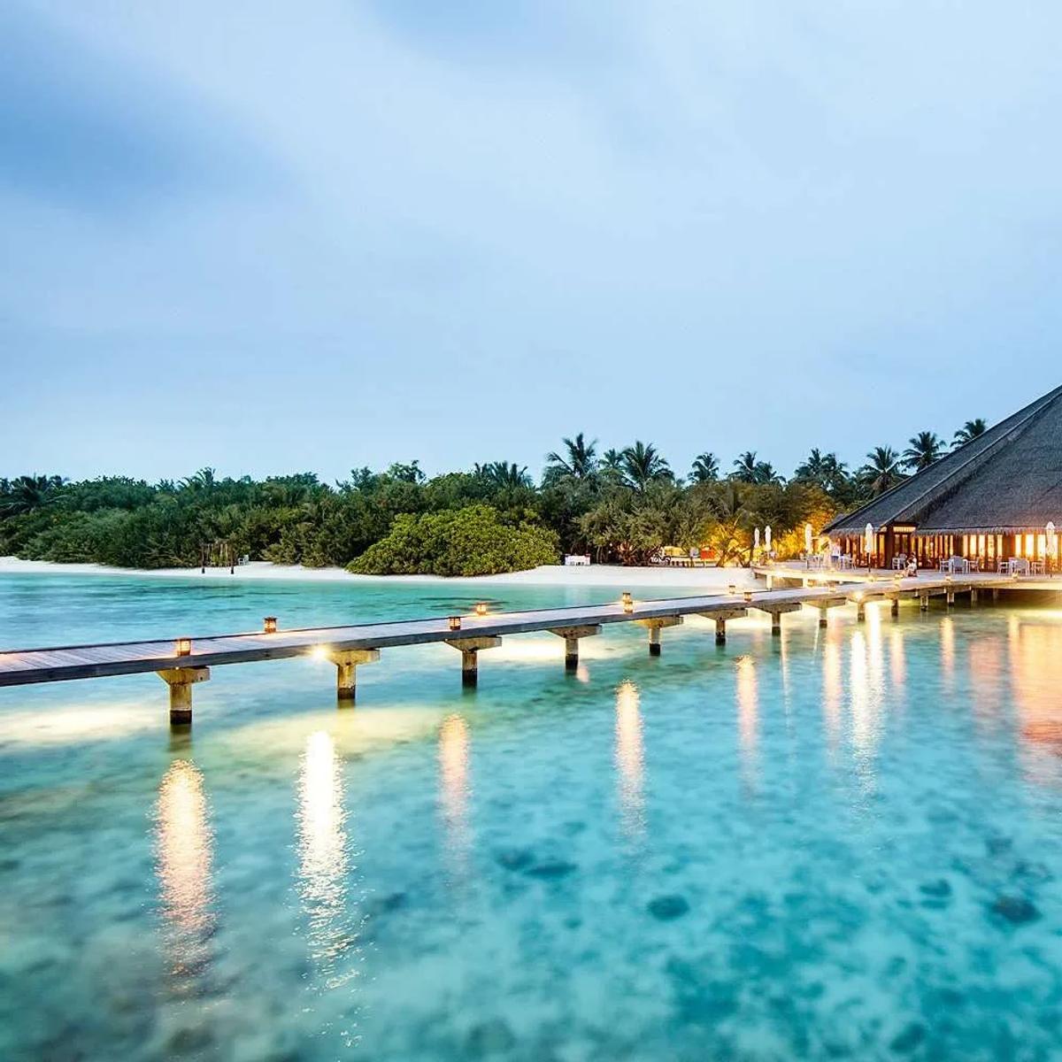 Matheefaru Restaurant lit at dusk, its jetty and thatched roof over the lagoon, Hideaway Beach Resort & Spa