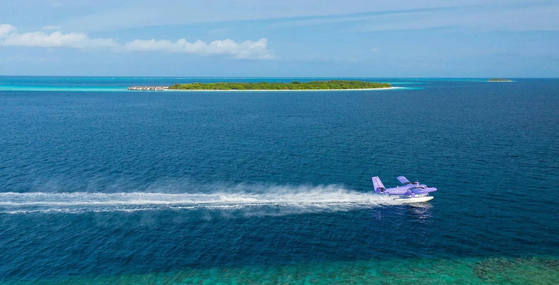 A Nautilus Maldives seaplane skimming low over open water, Thiladhoo Island visible in the distance.