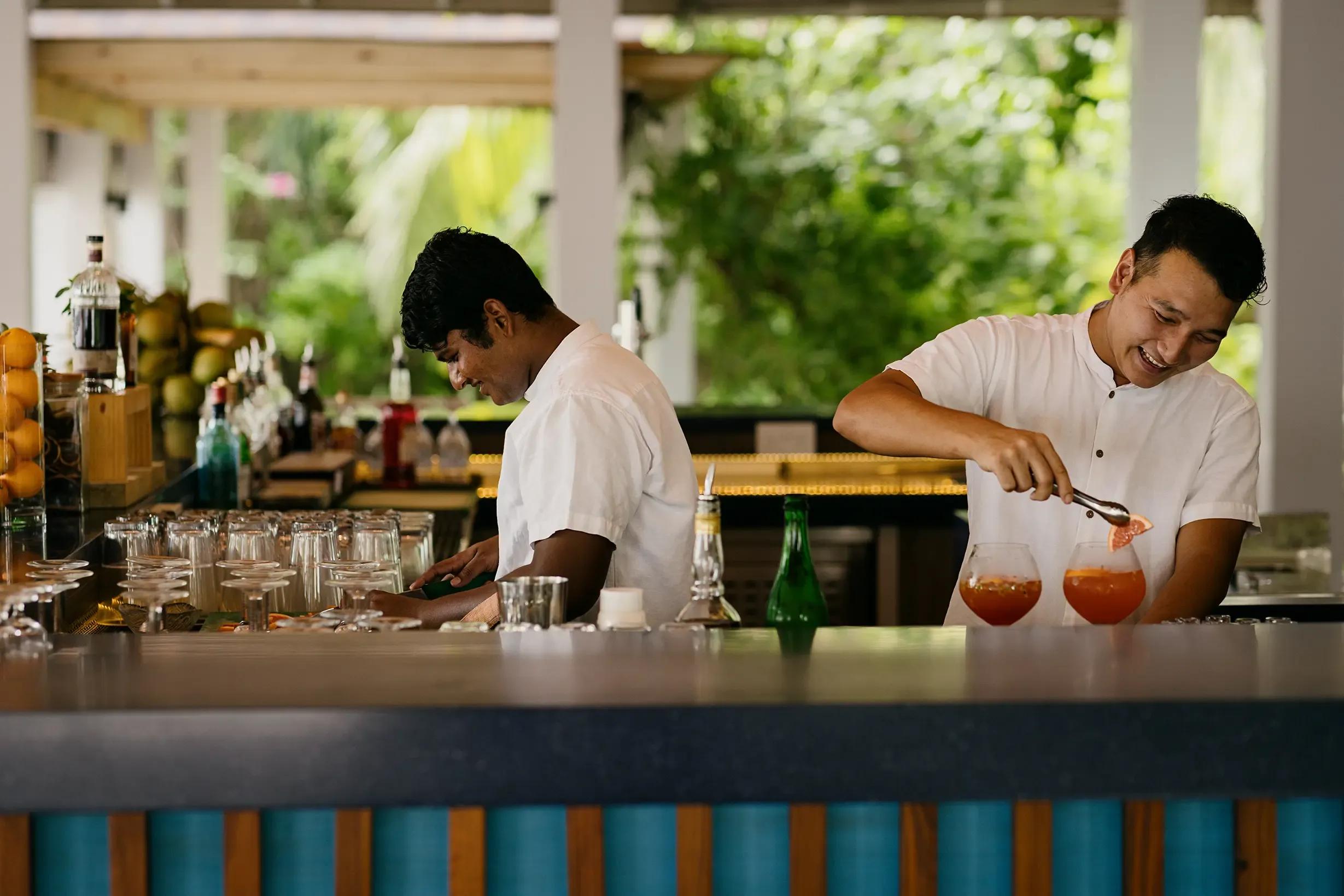 Two bartenders preparing cocktails behind the counter at East Bar, tropical planting behind them