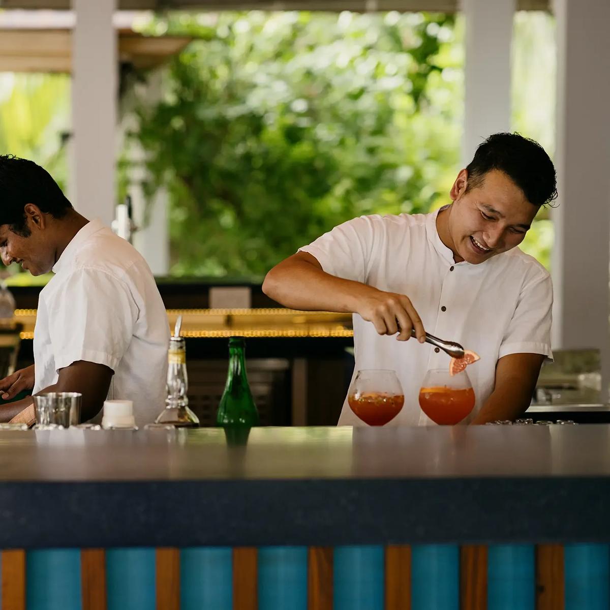 Two bartenders preparing cocktails behind the counter at East Bar, tropical planting behind them