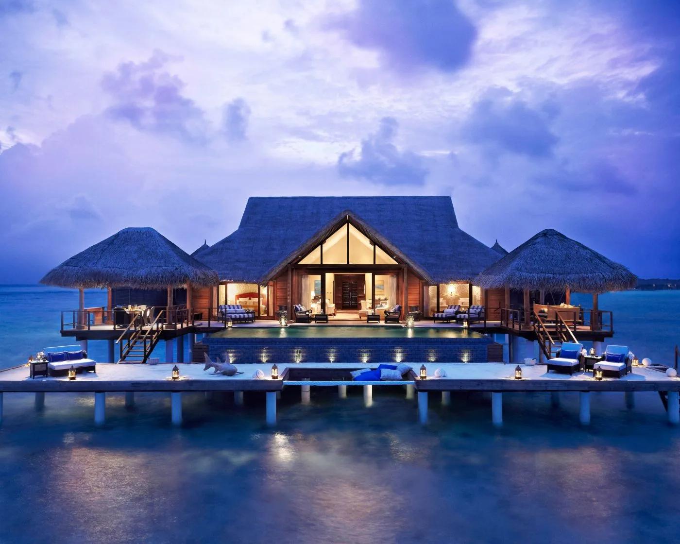 The Rehendhi Presidential Suite at dusk, its symmetrical thatched pavilions lit warmly over the lagoon