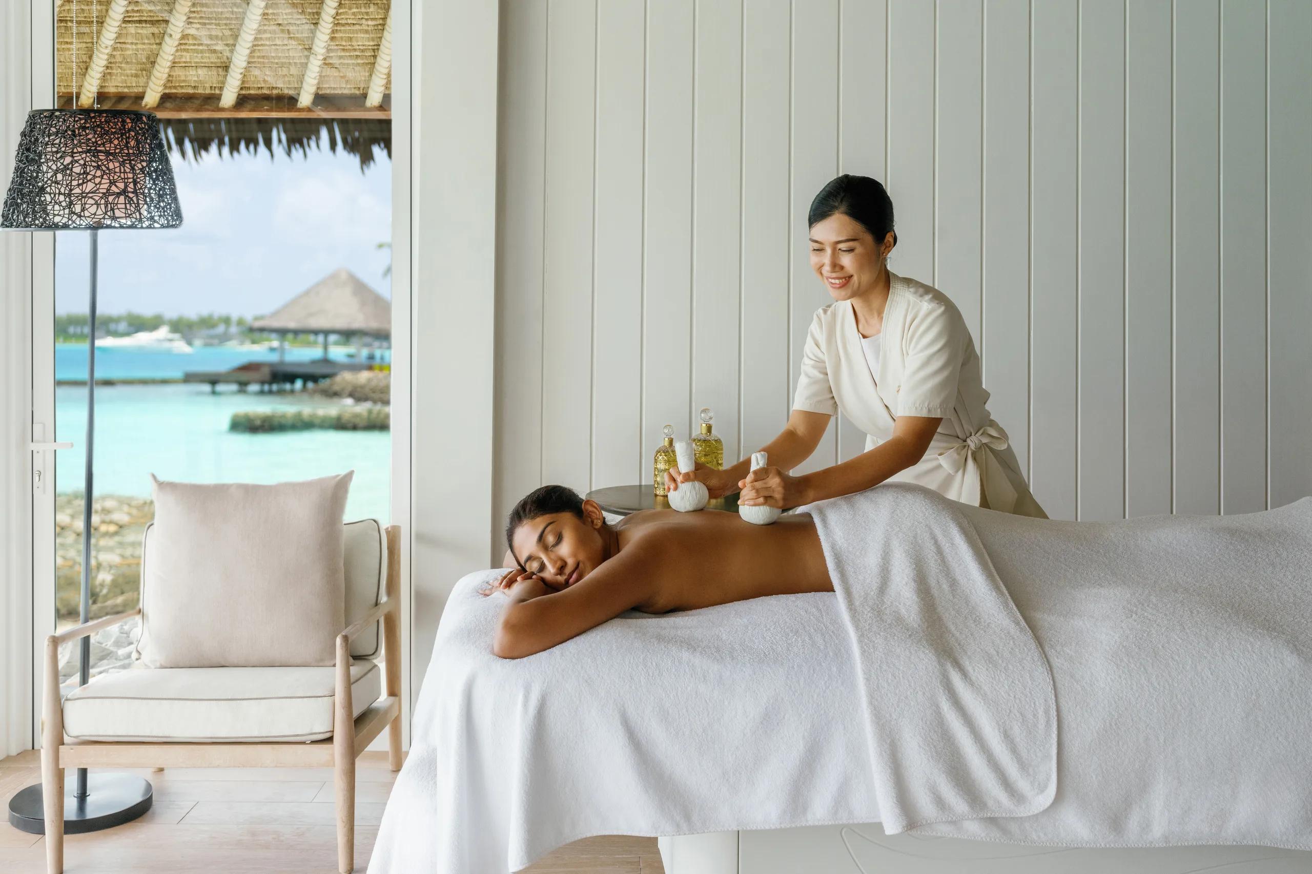 A therapist applies a herbal compress during a massage, the lagoon and a moored yacht visible through the window
