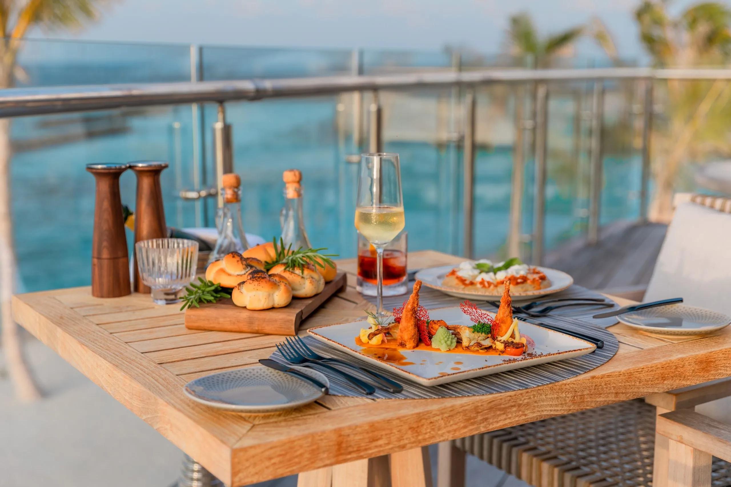 A table set for two on an ocean-view deck at dusk, with bread, a seafood starter and a glass of wine