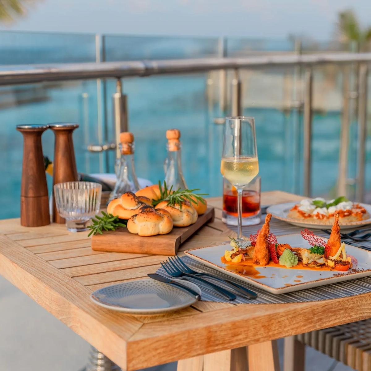 A table set for two on an ocean-view deck at dusk, with bread, a seafood starter and a glass of wine