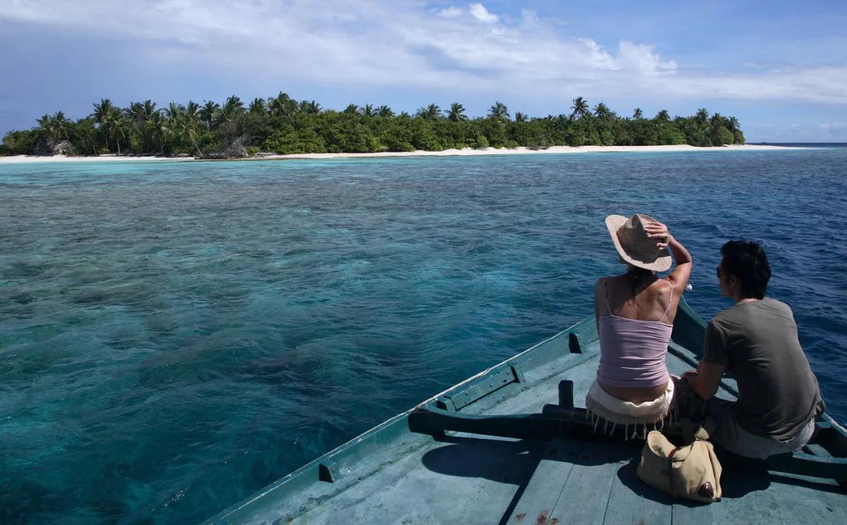 A couple seated at the bow of a traditional dhoni boat, sailing toward a palm-lined island