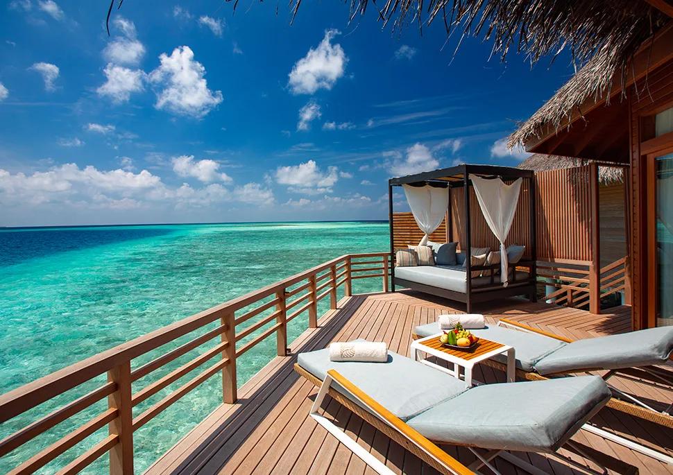 The deck of a Baros Water Villa, sun loungers and a shaded daybed looking straight out over the reef-lined lagoon