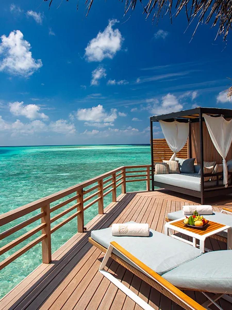The deck of a Baros Water Villa, sun loungers and a shaded daybed looking straight out over the reef-lined lagoon