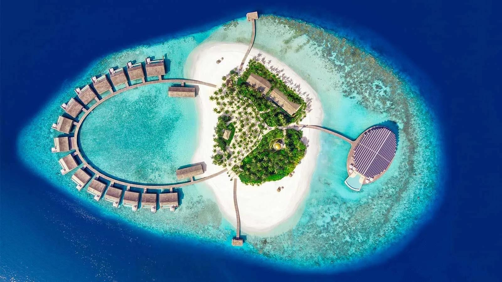 A direct top-down aerial of Kudadoo's island, its horseshoe of overwater residences curving around the lagoon beside the solar-roofed Retreat