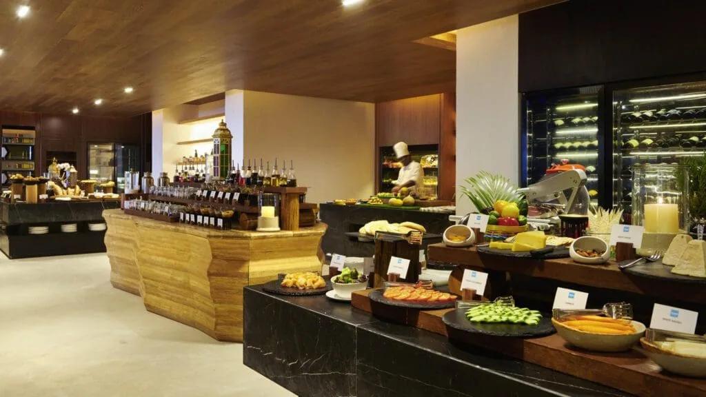 A chef preparing dishes at an open buffet counter, with a glass wine cooler and small plates of food arranged in the foreground