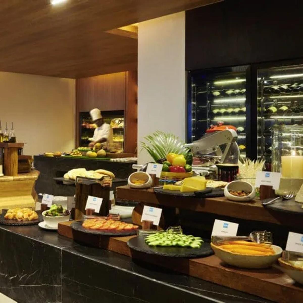 A chef preparing dishes at an open buffet counter, with a glass wine cooler and small plates of food arranged in the foreground