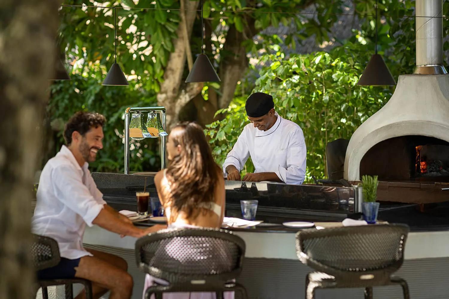 Guests seated at Fogliani's open-air kitchen counter as a chef prepares food beside the wood-fired pizza oven