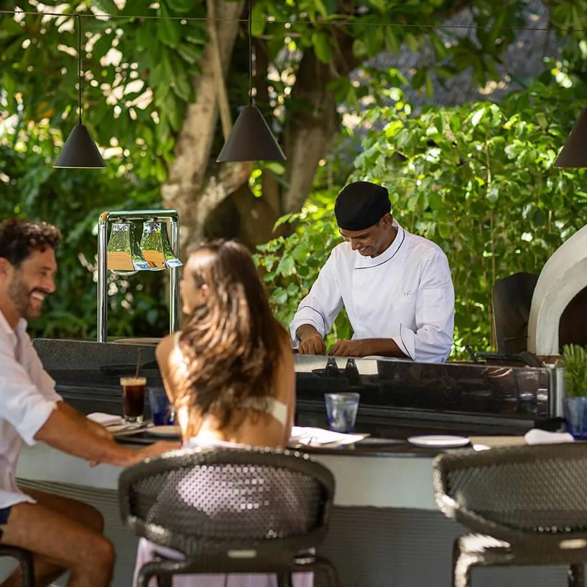 Guests seated at Fogliani's open-air kitchen counter as a chef prepares food beside the wood-fired pizza oven