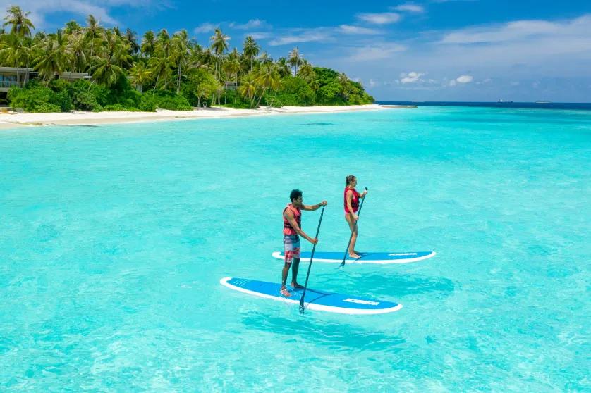 Two guests paddleboarding across a shallow turquoise lagoon, the island's palm-lined shore behind them