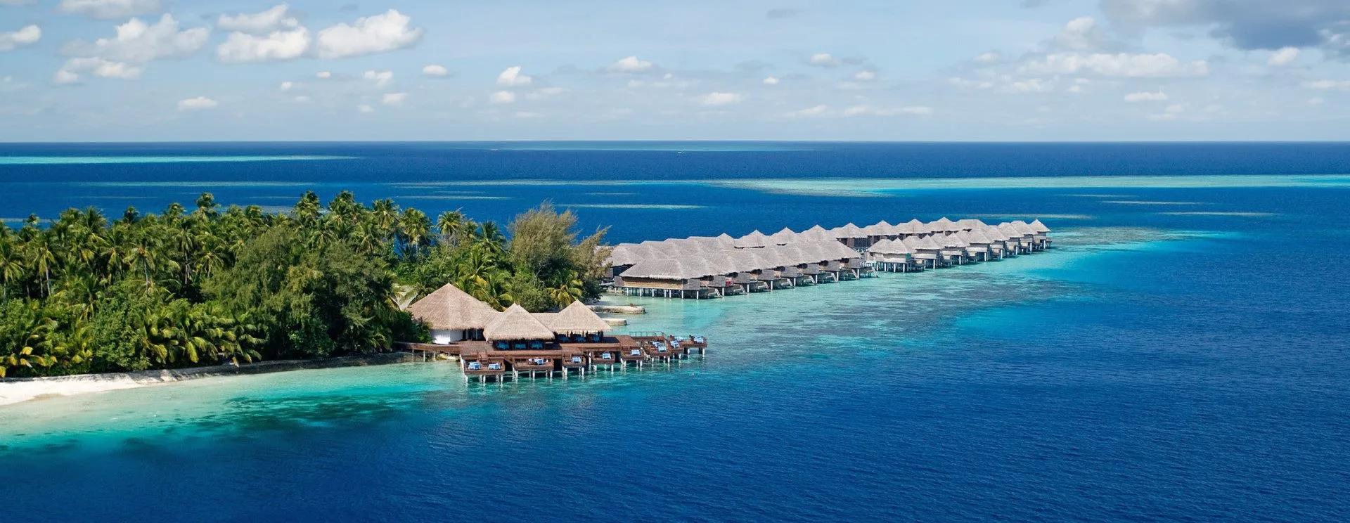 Coco Bodu Hithi's overwater villas curving along the reef edge beside the island's palm-covered shoreline