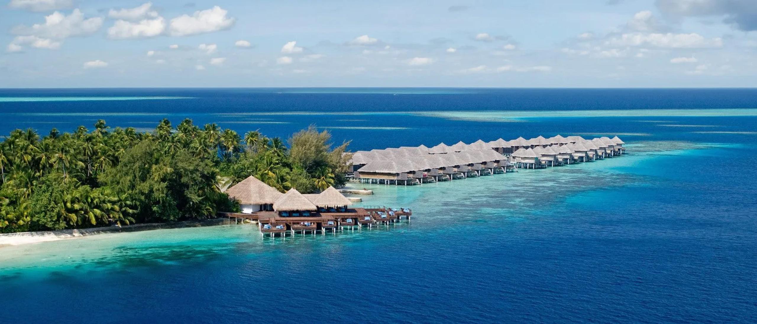 Coco Bodu Hithi's overwater villas curving along the reef edge beside the island's palm-covered shoreline