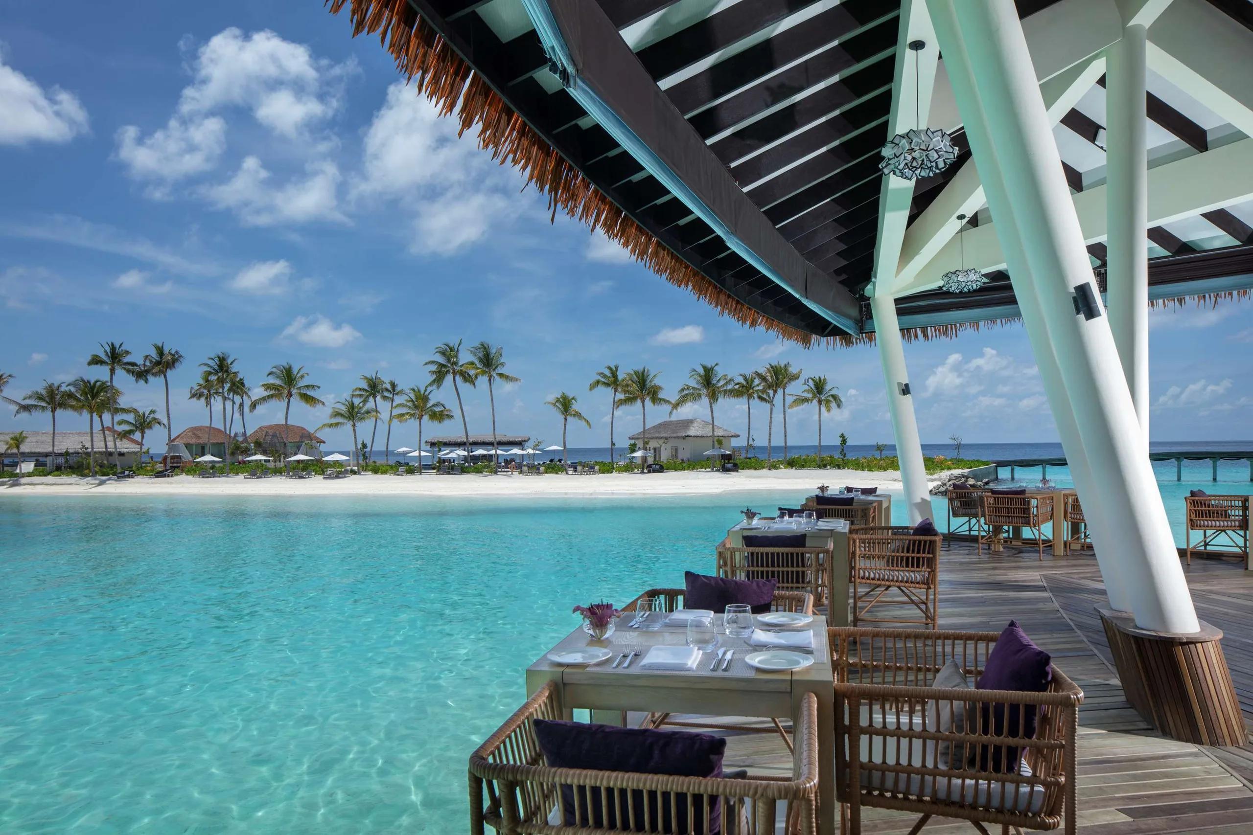 Raha's overwater dining deck at Radisson Blu Resort Maldives, tables set above a turquoise lagoon with a sandbar and palm trees beyond