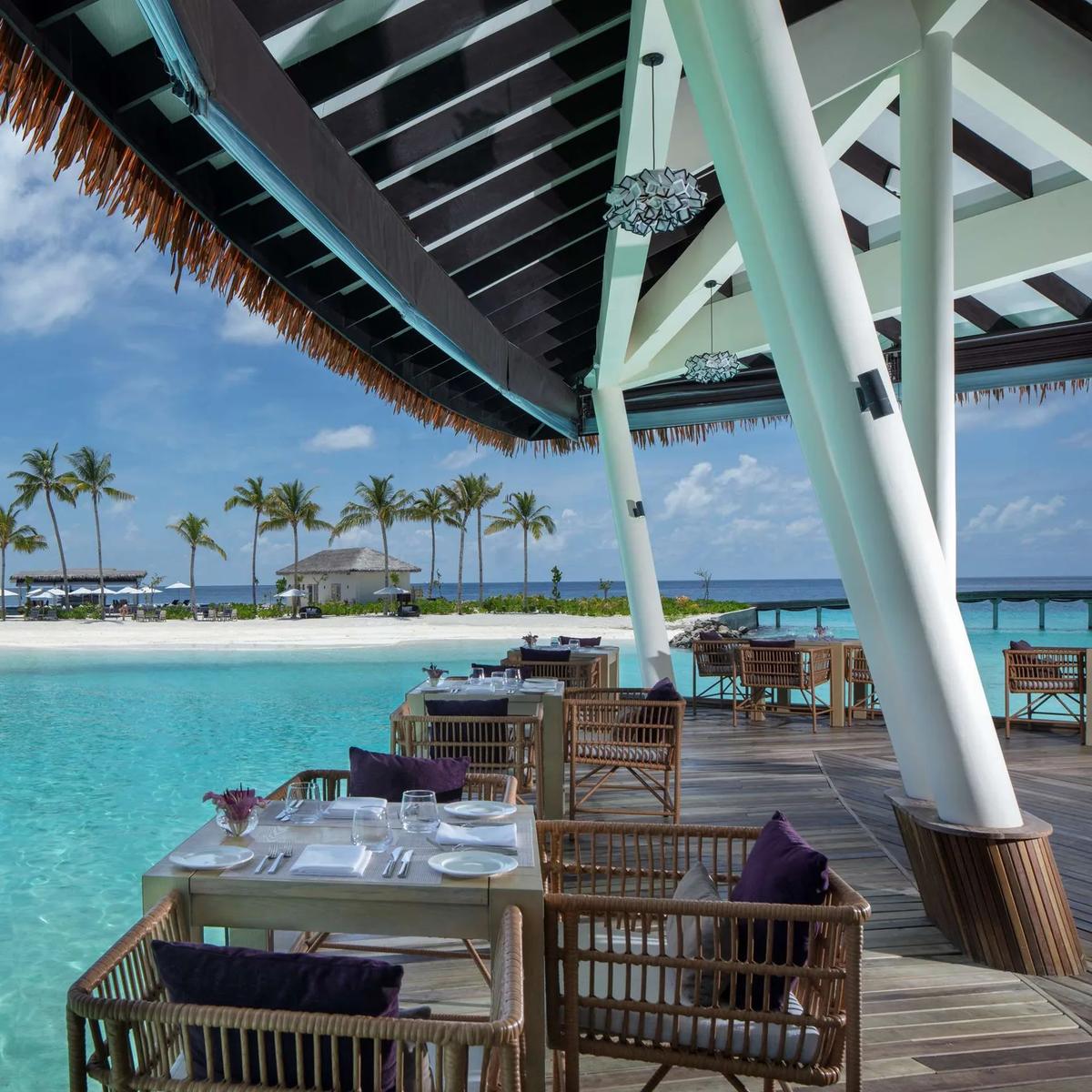 Raha's overwater dining deck at Radisson Blu Resort Maldives, tables set above a turquoise lagoon with a sandbar and palm trees beyond