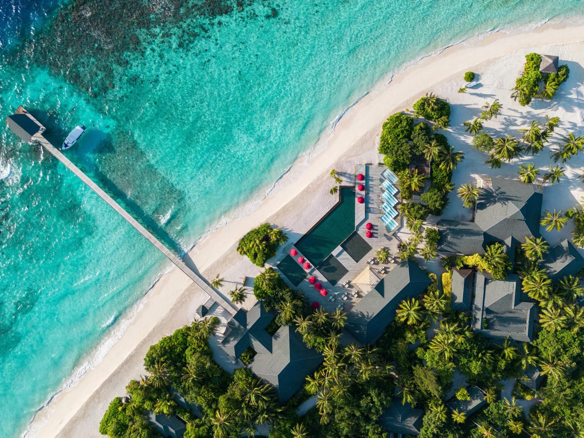 Closer aerial view of Havodda's point, with a wooden jetty reaching into turquoise water, a moored speedboat, and a thatched-roof pool deck with red umbrellas and loungers among the palms