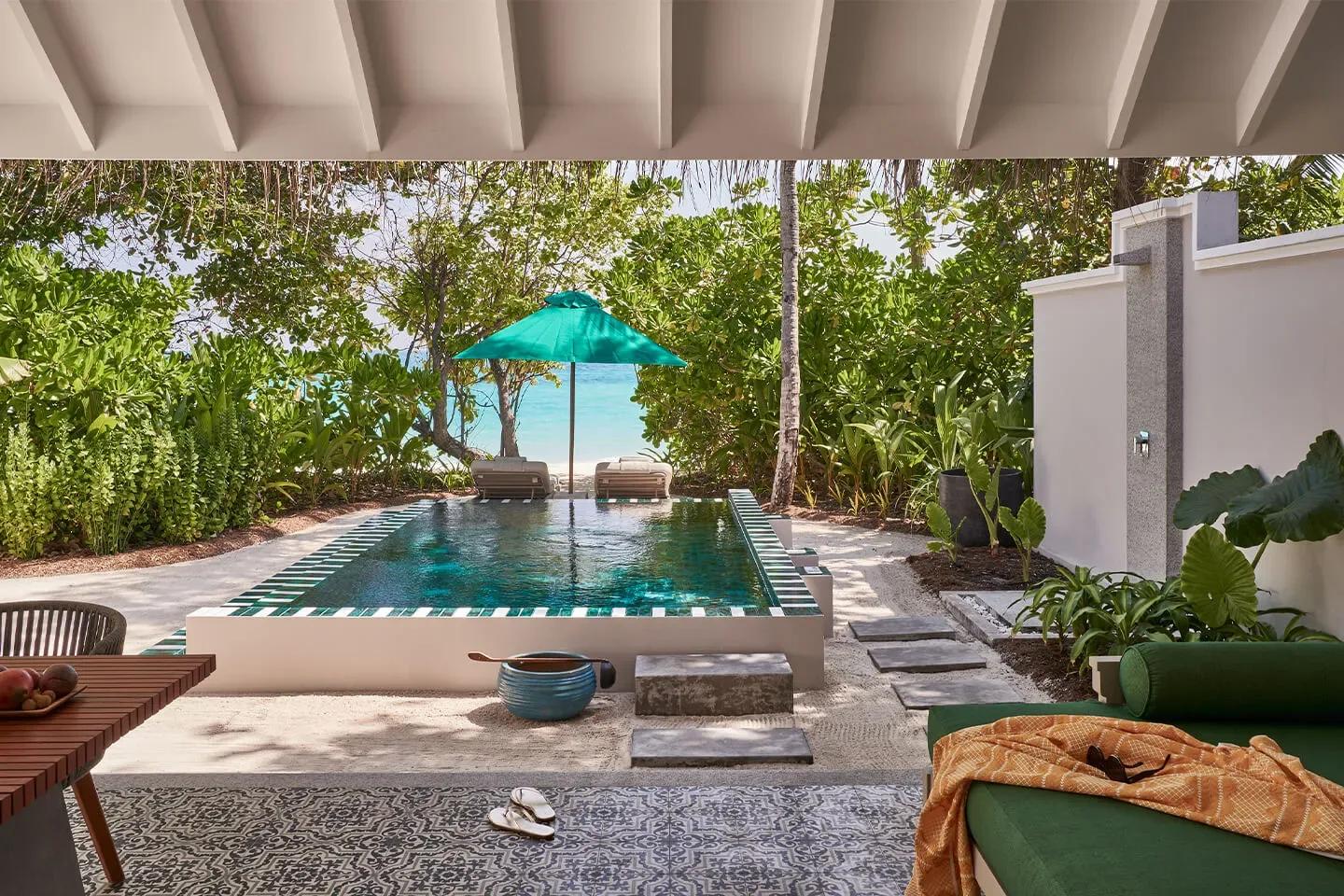 A Beach Pool Villa's private mosaic-tiled pool under a leaning palm and a teal umbrella, the beach visible through the greenery beyond