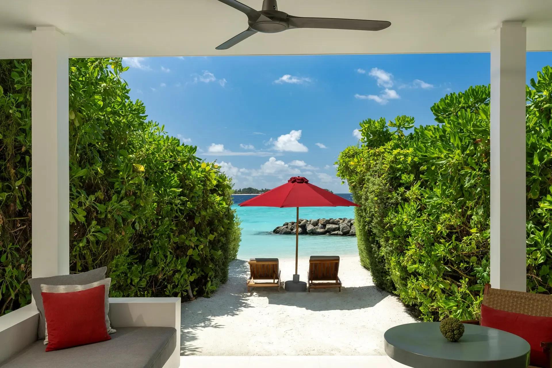 A shaded terrace opening onto a sandy path lined with palms, leading to loungers on the beach