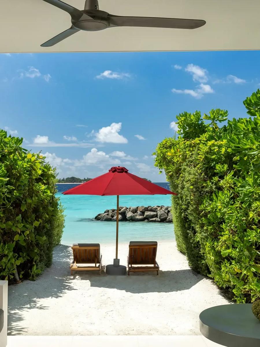 A shaded terrace opening onto a sandy path lined with palms, leading to loungers on the beach