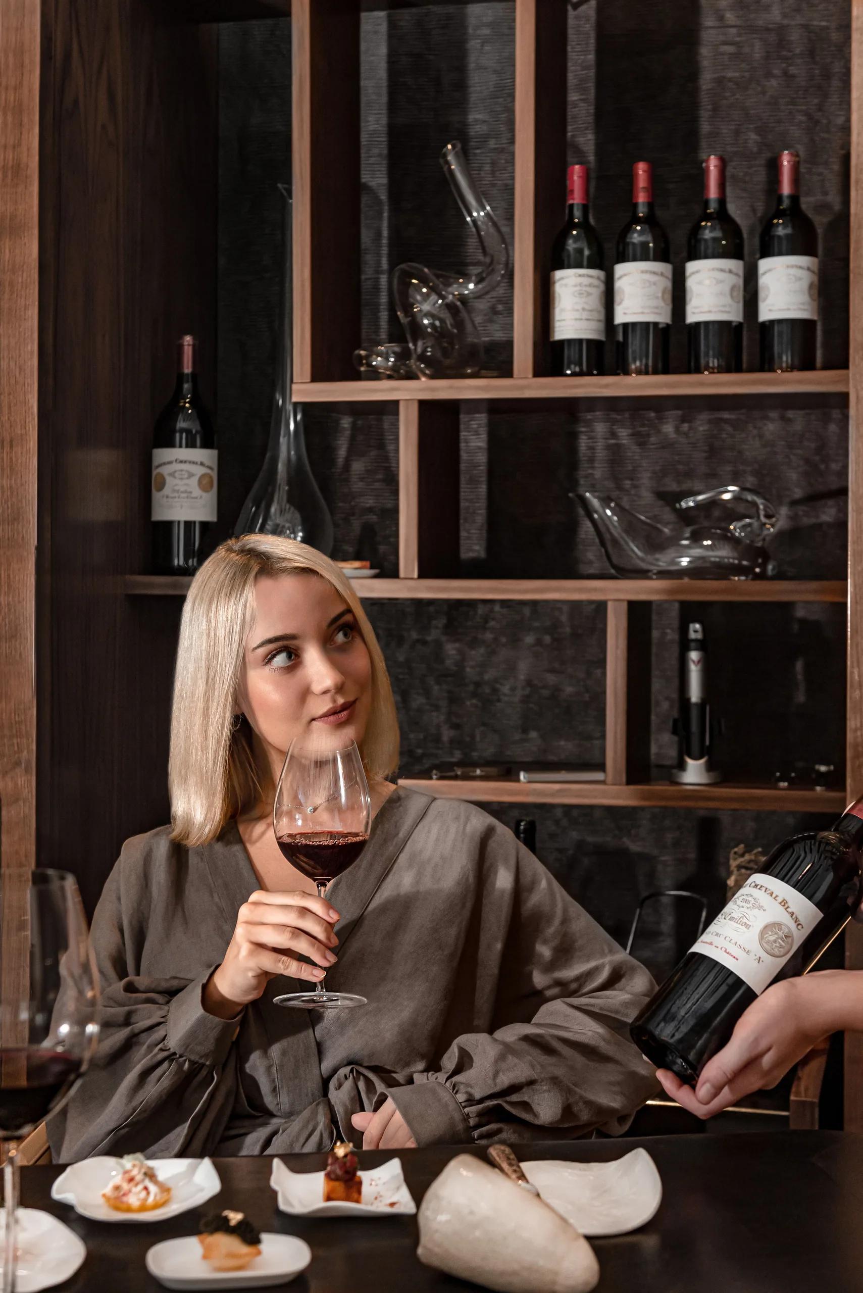 A guest tastes red wine in the Le 1947 wine cellar, shelves of Château Cheval Blanc bottles behind her