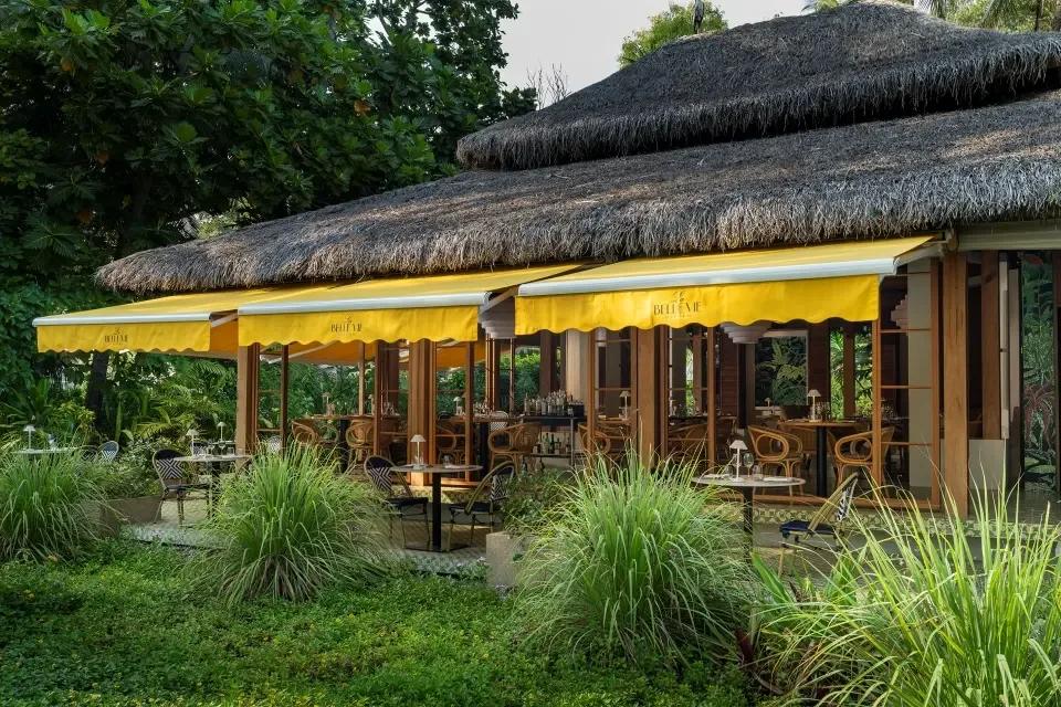 La Belle Vie's thatched-roof exterior with yellow awnings over open-air tables, set in a garden of tall grasses