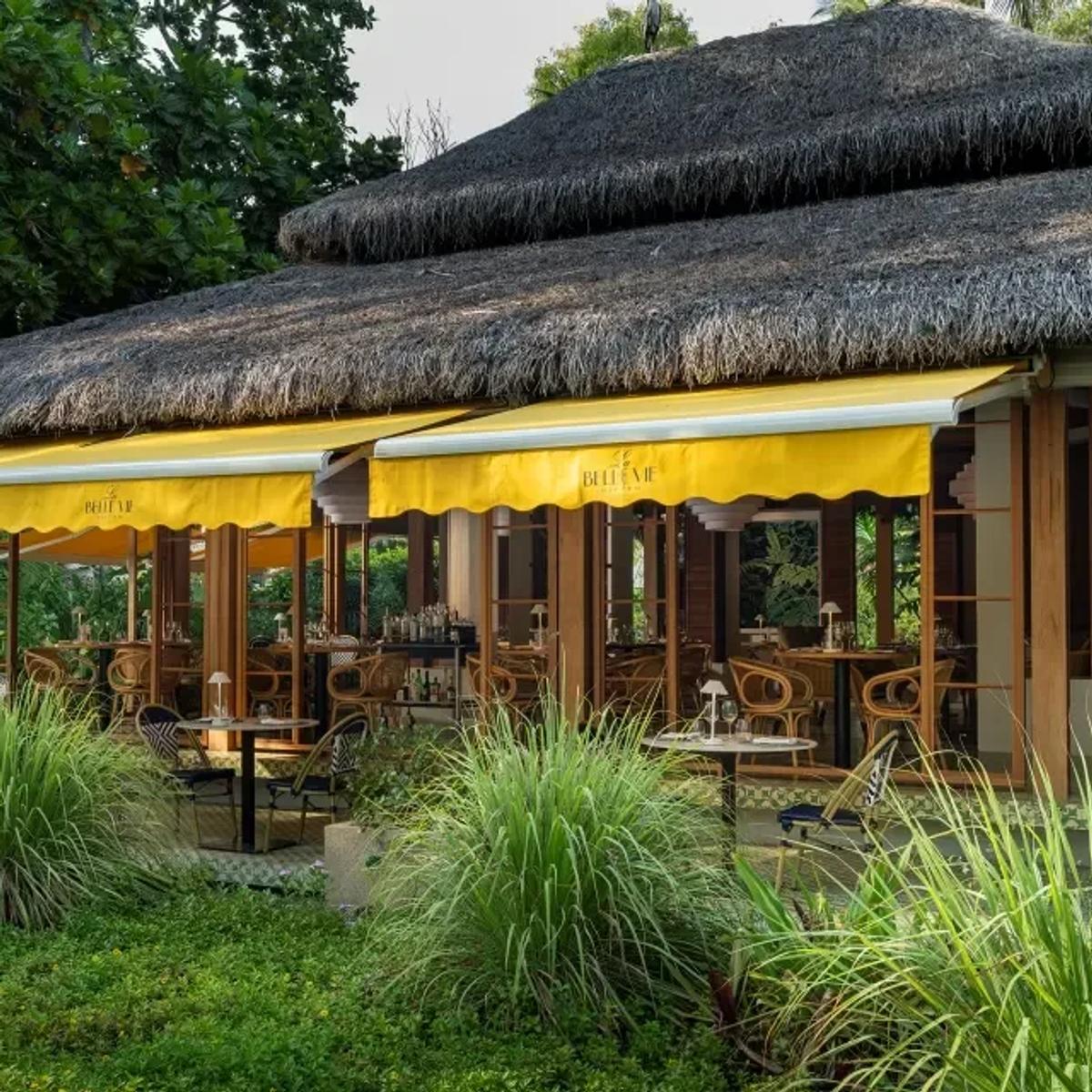 La Belle Vie's thatched-roof exterior with yellow awnings over open-air tables, set in a garden of tall grasses