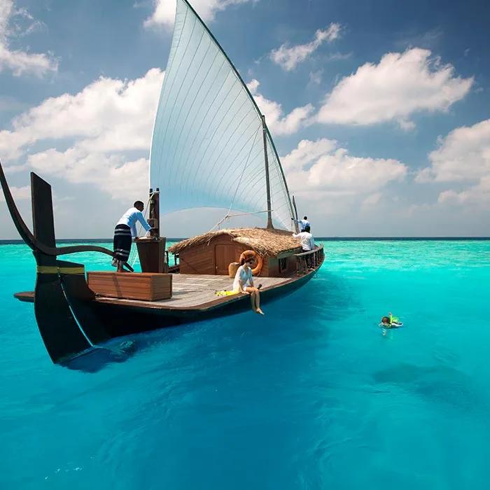 The traditional sailing dhoni Nooma at anchor with its sail raised, a guest resting on deck and a snorkeller in the water nearby