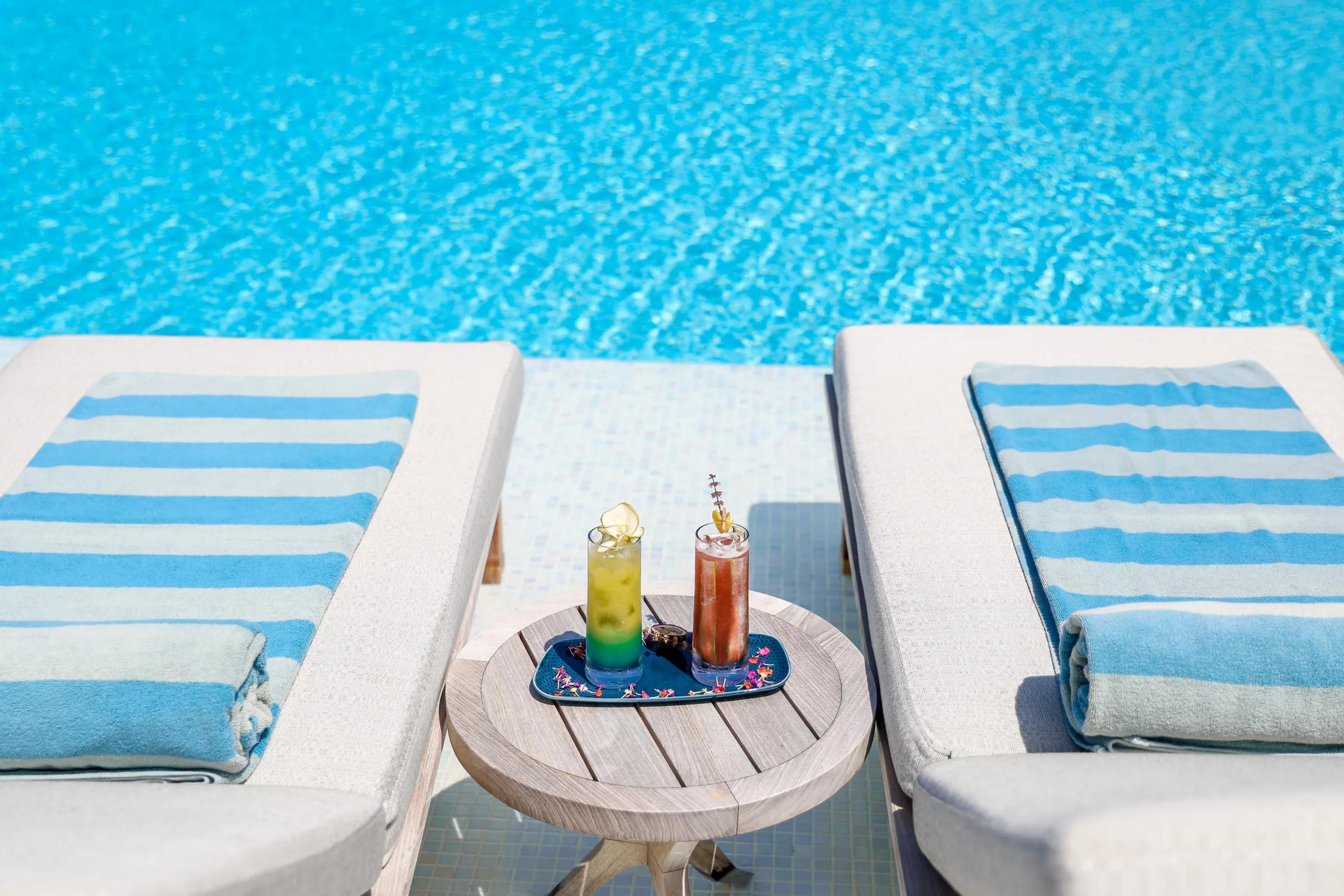 Two poolside cocktails on a tray between a pair of striped sun loungers