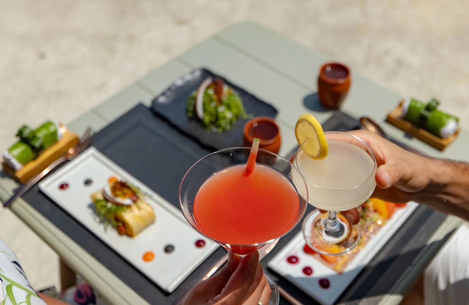 Two cocktails held over a table of small plates, seen from above on a sunlit terrace
