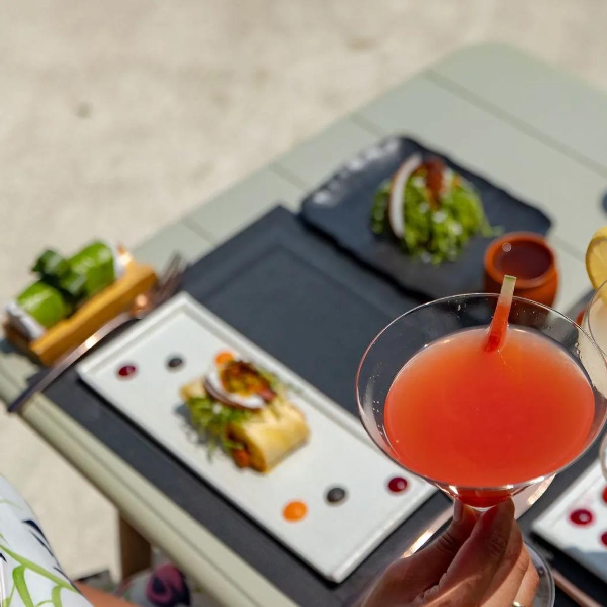 Two cocktails held over a table of small plates, seen from above on a sunlit terrace