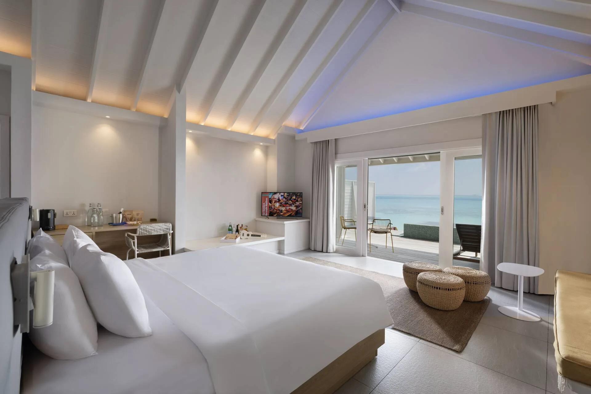 The bedroom of an Ocean Overwater Villa at The Standard, a television and deck doors opening onto the lagoon visible beyond the bed