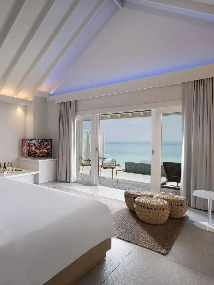 The bedroom of an Ocean Overwater Villa at The Standard, a television and deck doors opening onto the lagoon visible beyond the bed