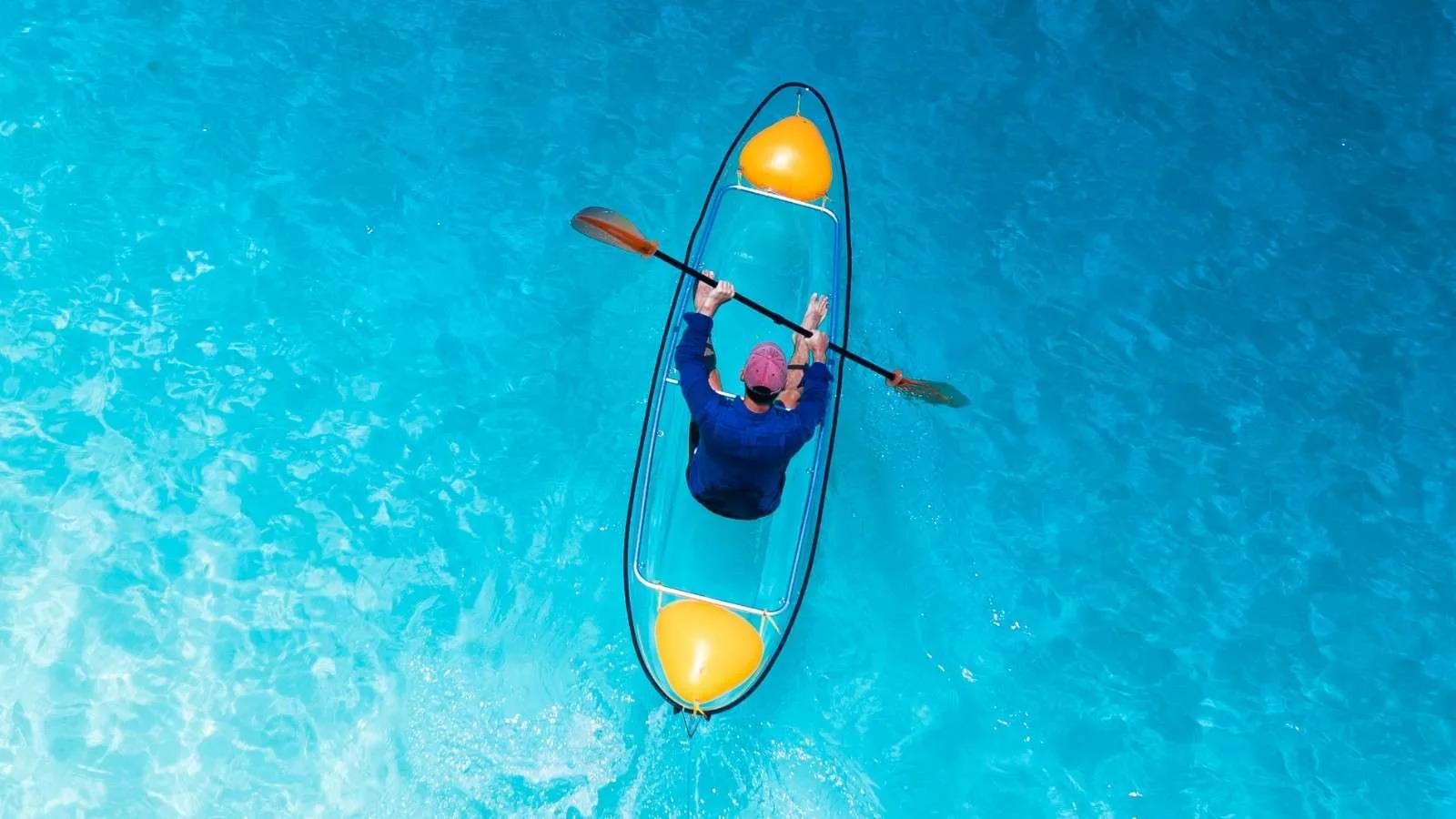 An overhead view of a guest paddling a glass-bottom kayak through clear turquoise water at Jawakara