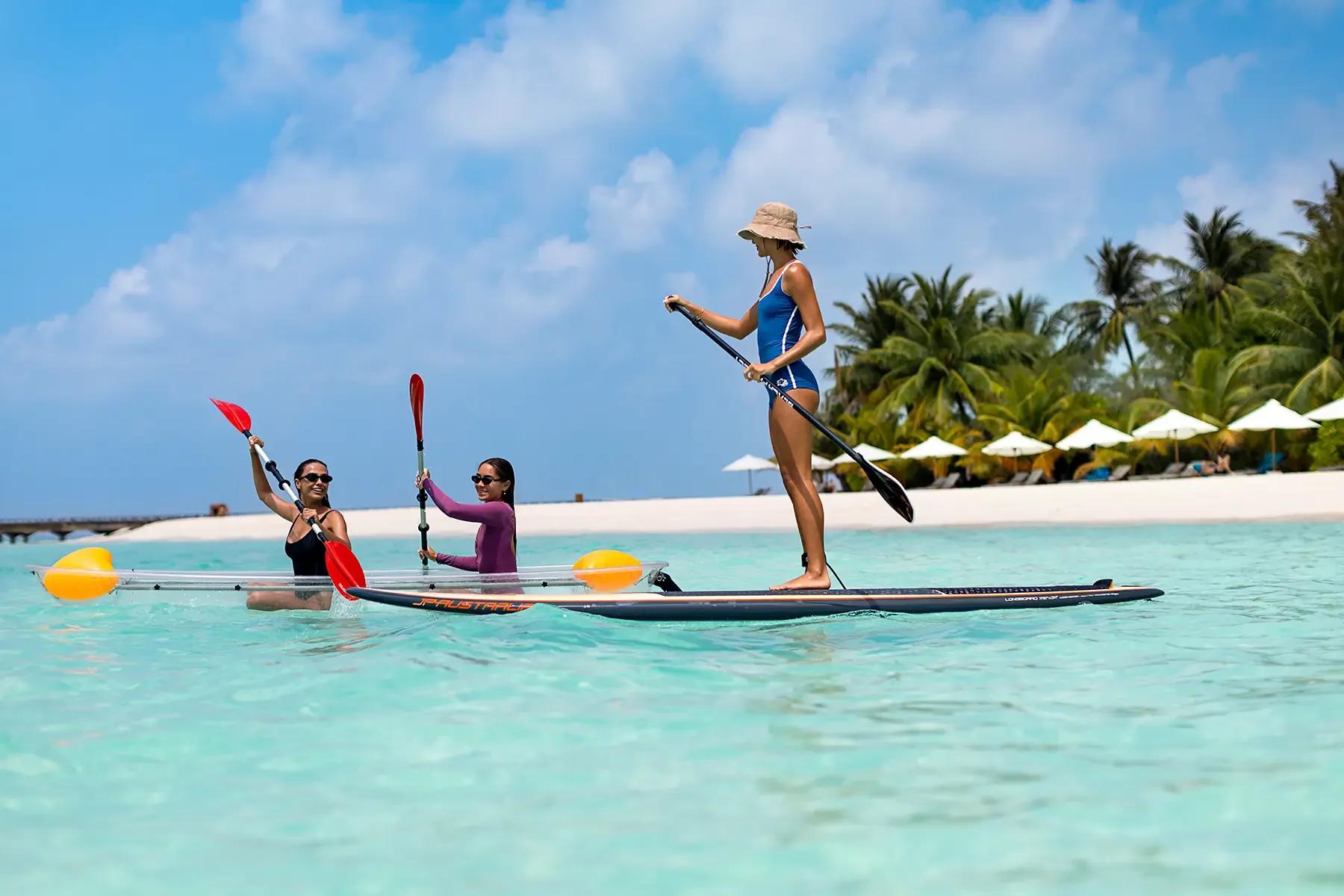Three guests on a stand-up paddleboard and a clear-hulled kayak in shallow water near the beach
