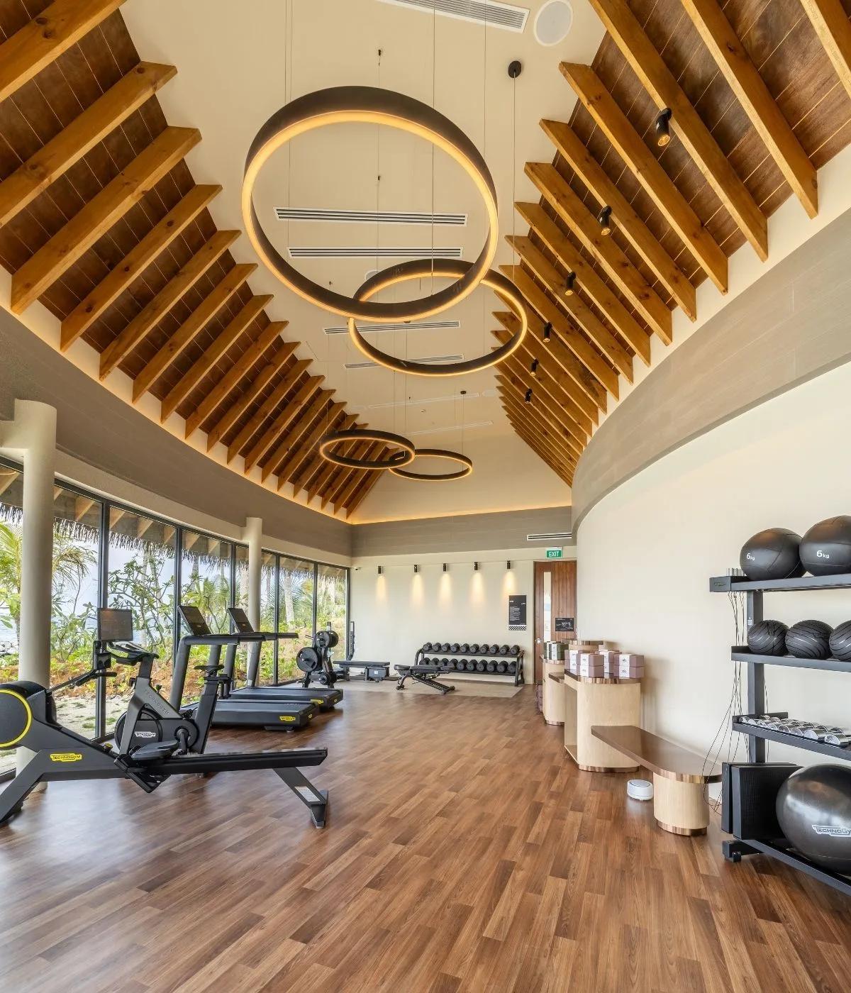 The gym at SO/ Maldives, its vaulted timber roof hung with ring light fixtures above rows of cardio and strength equipment