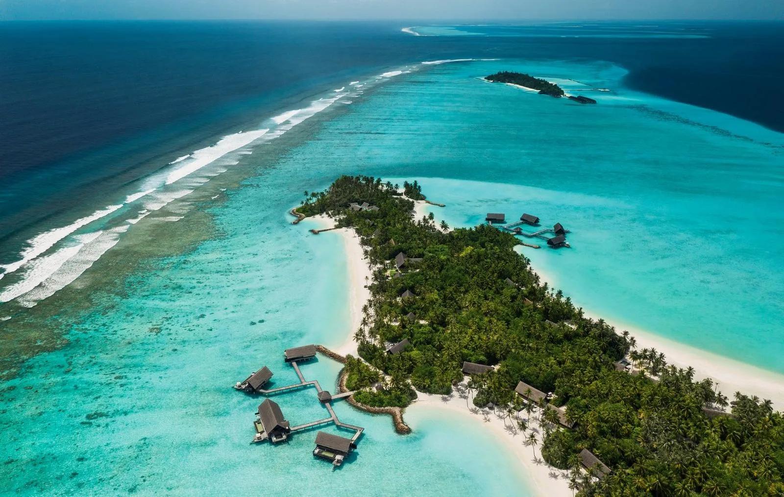 Aerial view of One&Only Reethi Rah's island in North Malé Atoll, water villas reaching into the lagoon on stilted walkways and a line of surf breaking on the outer reef