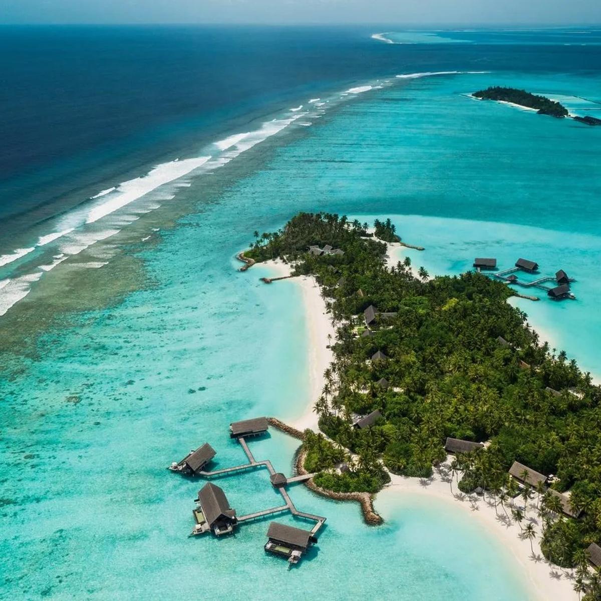 Aerial view of One&Only Reethi Rah's island in North Malé Atoll, water villas reaching into the lagoon on stilted walkways and a line of surf breaking on the outer reef