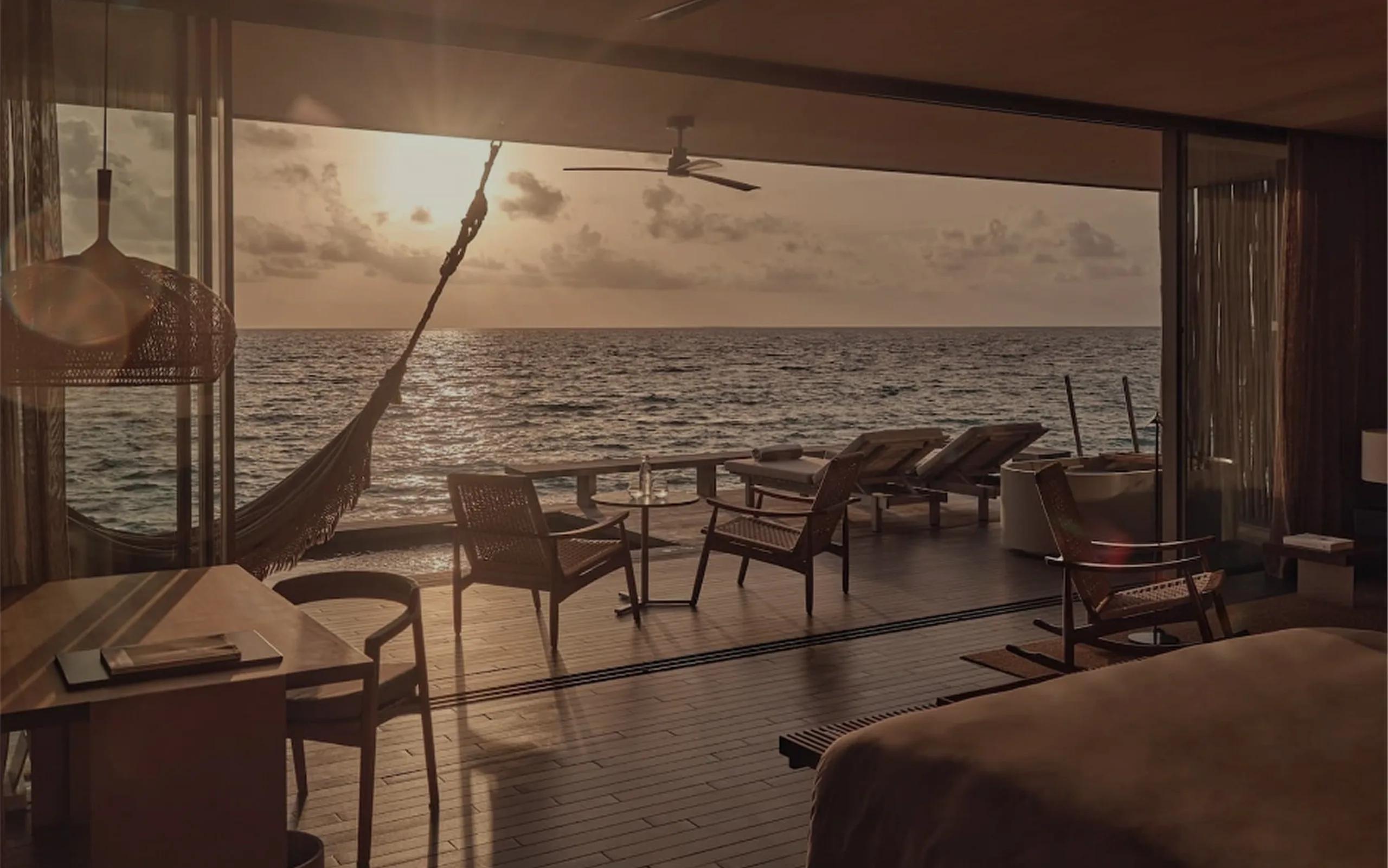 A hammock strung above the deck of a One-Bedroom Sunset Water Pool Villa at dusk, chairs and a table set out over the open ocean