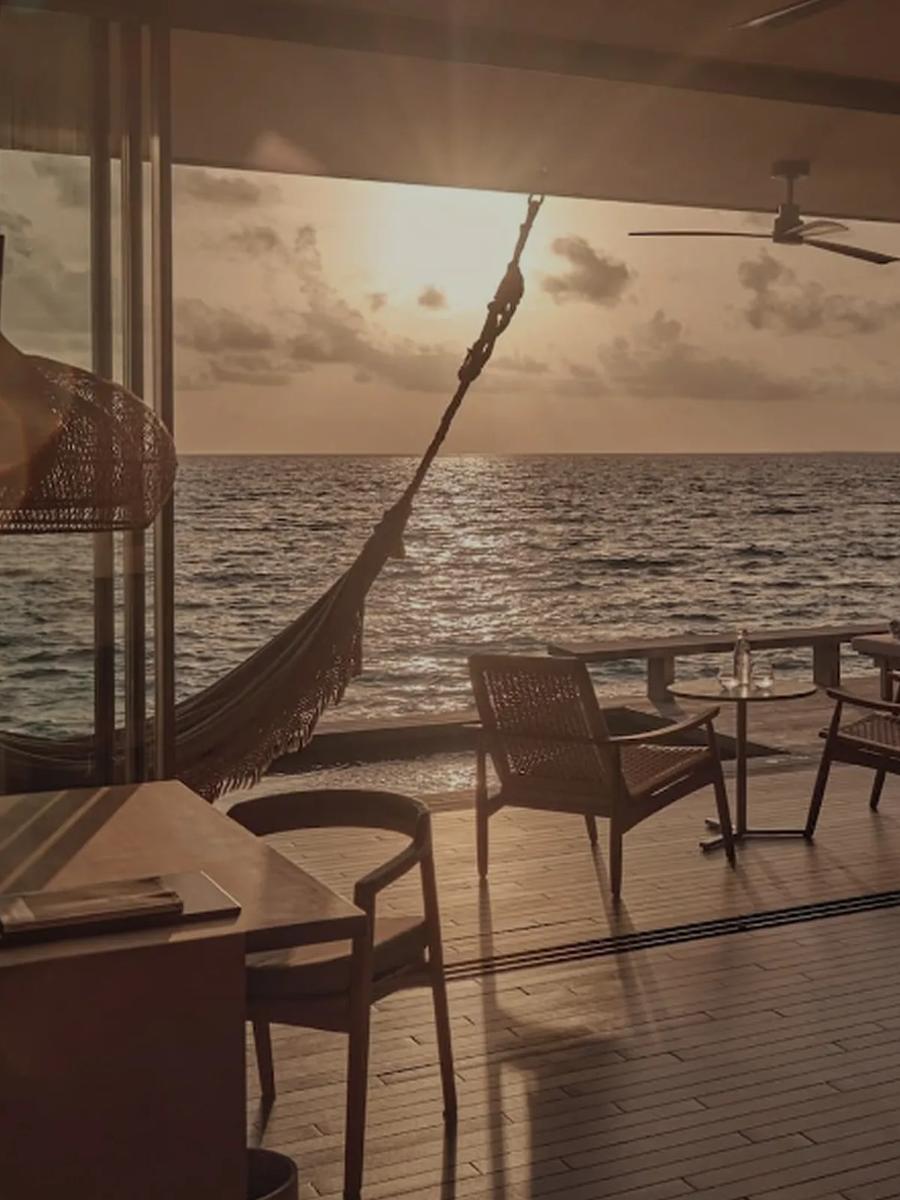 A hammock strung above the deck of a One-Bedroom Sunset Water Pool Villa at dusk, chairs and a table set out over the open ocean