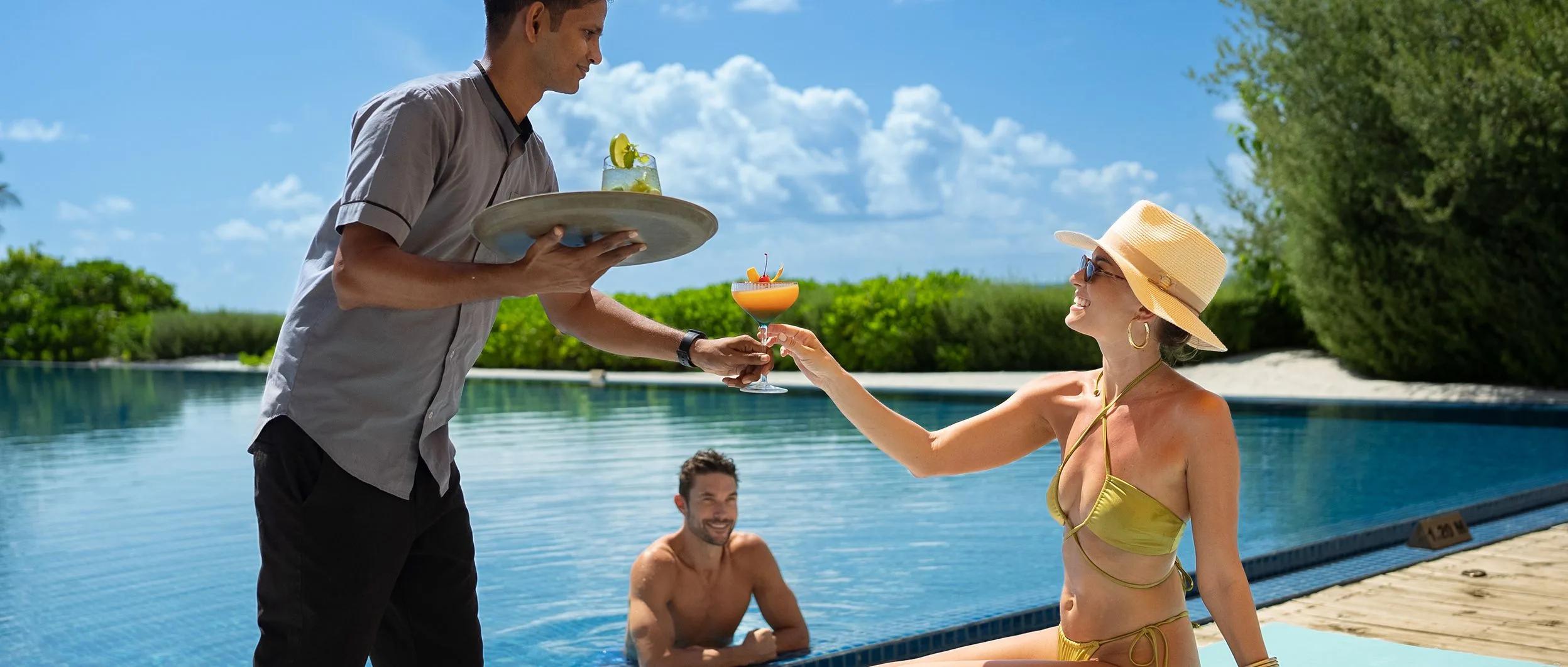 A bartender hands a cocktail to a guest at the main pool's edge, The Liquid's bar behind