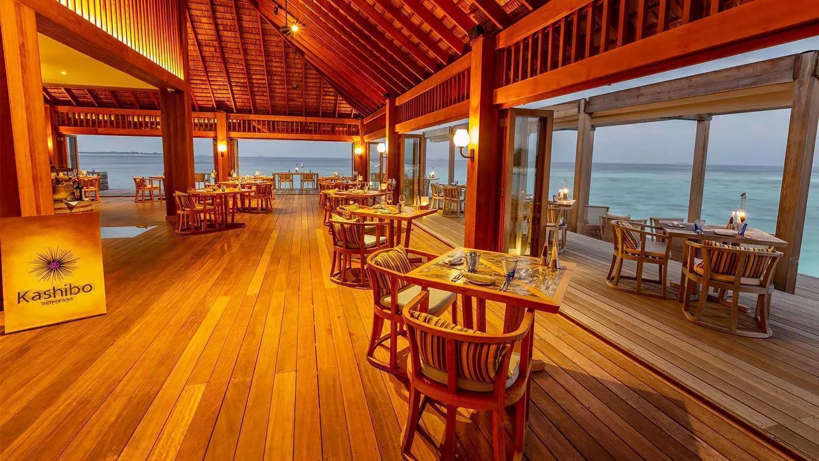 Kashibo's overwater dining room at dusk, tables set along glass walls facing the lagoon