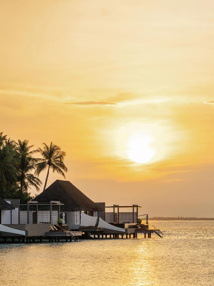 Overwater Water Villas on stilts above the lagoon at sunset, palm trees silhouetted along the walkway
