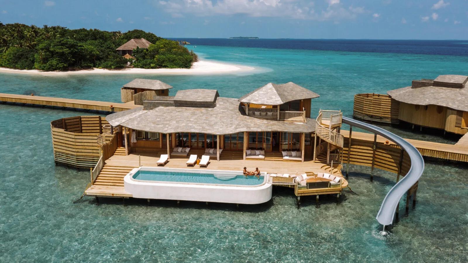 An overwater villa's curved infinity pool built out over the lagoon, with a slide down into the water and two guests relaxing in the pool